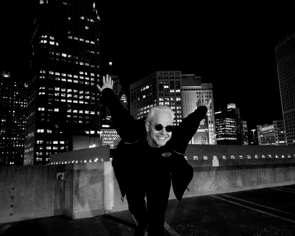 Adam leaning into the camera with arms extended behind him. He is grinning awkwardly. Cityscape in the background.