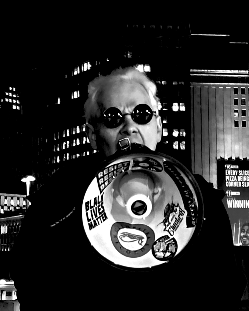 Adam yelling into a megaphone. Cityscape in the background.