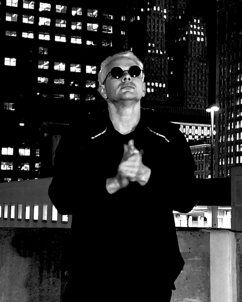 Adam is rubbing his hands together and looking up and away. Cityscape in the background.